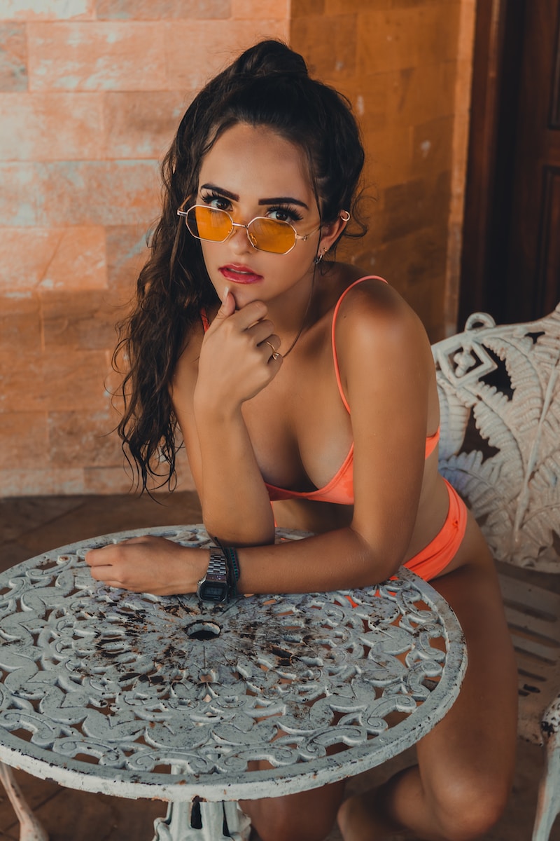 woman wearing orange bikini leaning forward on patio table with right hand touching her chin